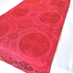 May include: Red fabric with a repeating pattern of ornate circles containing crosses. The fabric has a subtle sheen.