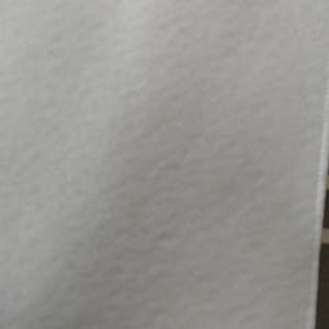May include: A close-up of a white, textured fabric. The material appears soft and slightly uneven, with a subtle, light gray hue. The edge of the fabric is visible on the right side, showing a clean, finished seam.