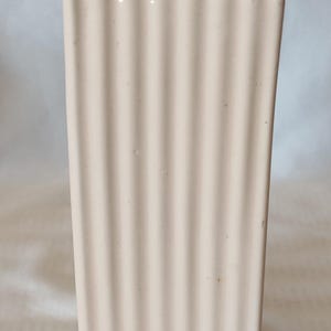 May include: A tall, rectangular ceramic vase in a pale cream color. The exterior features a vertical ribbed texture that runs from the top rim to the base. The vase has a hollow center and a flat, square-shaped opening at the top.