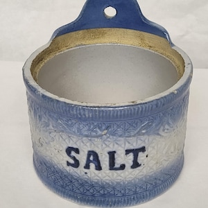 Könnte beinhalten: Ein zylindrisches Salzgefäß aus Keramik in Blau und Weiß mit einem strukturierten Rautenmuster auf der Außenseite. Das Wort SALT ist in dunkelblauen Buchstaben auf der Vorderseite aufgedruckt, und oben befindet sich eine Lasche mit Loch.