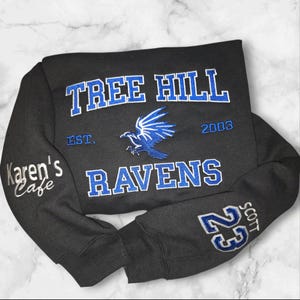 May include: Black sweatshirt with blue and white embroidered text and graphics. The front features "TREE HILL RAVENS" with a raven graphic and "EST. 2003." The left sleeve has "Karen's Cafe" and the right sleeve has "23 SCOTT."