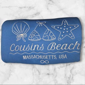 May include: A blue beach towel with white embroidered designs of a seashell, bikini top, and starfish. The text "Cousins Beach" and "Massachusetts, USA" are also embroidered on the towel.