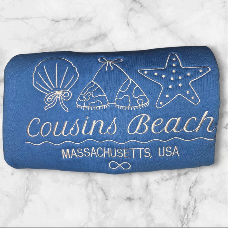 May include: A blue beach towel with white embroidered designs of a seashell, bikini top, and starfish. The text "Cousins Beach" and "Massachusetts, USA" are also embroidered on the towel.