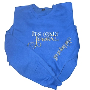 May include: A royal blue sweatshirt with the text "IT'S ONLY forever..." in white and gold script on the front. The sleeve has the text "Not long at all" in gold script. The sweatshirt is made of a soft material.