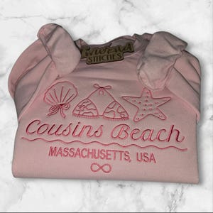 May include: A light pink sweatshirt with embroidered beach-themed designs, including a seashell, bikini top, and starfish. The text "Cousins Beach" and "Massachusetts, USA" are embroidered below the designs. The sweatshirt also has the "Emerald Stitches" tag.