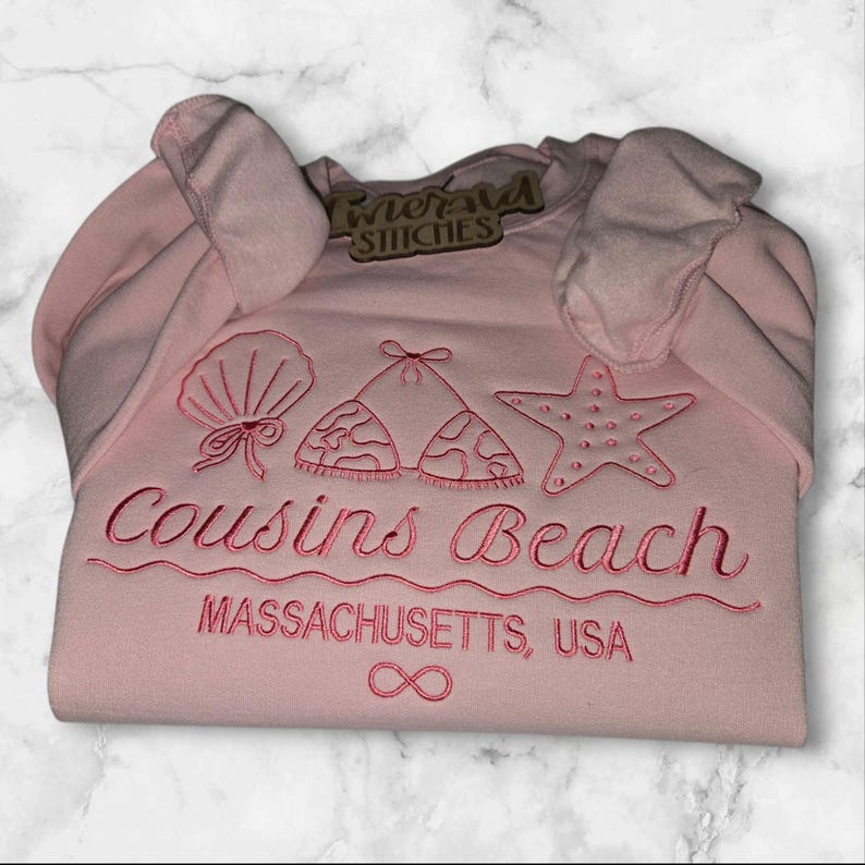 May include: A light pink sweatshirt with embroidered beach-themed designs, including a seashell, bikini top, and starfish. The text "Cousins Beach" and "Massachusetts, USA" are embroidered below the designs. The sweatshirt also has the "Emerald Stitches" tag.