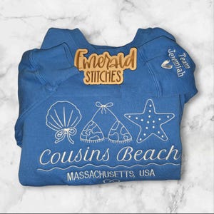 May include: A blue sweatshirt with white embroidered beach-themed designs, including a seashell, bikini tops, and a starfish. The text "Cousins Beach Massachusetts, USA" is embroidered below the graphics. A wooden sign that says "Emerald Stitches" is on top.