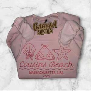 May include: A light pink sweatshirt with embroidered beach-themed designs, including a seashell, bikini top, and starfish. The text "Cousins Beach, Massachusetts, USA" is embroidered below the designs. A wooden tag with the text "Emerald Stitches" is attached.