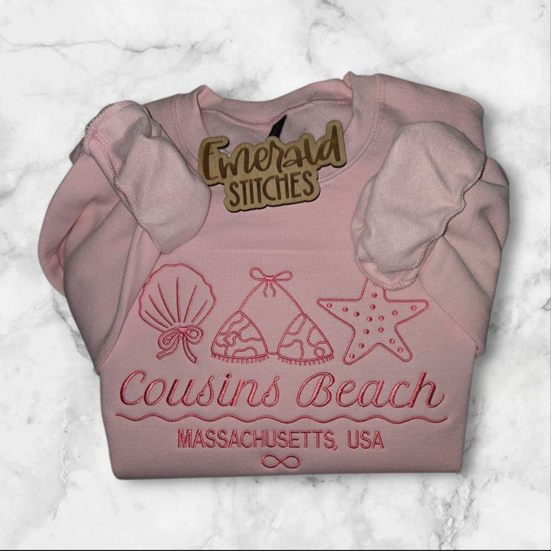 May include: A light pink sweatshirt with embroidered beach-themed designs, including a seashell, bikini top, and starfish. The text "Cousins Beach, Massachusetts, USA" is embroidered below the designs. A wooden tag with the text "Emerald Stitches" is attached.