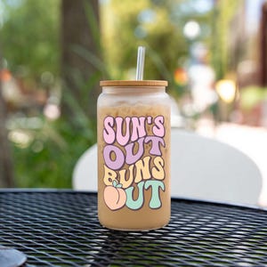 May include: A clear glass tumbler with a bamboo lid and a straw, filled with an iced coffee drink. The tumbler features the text "SUN'S OUT BUNS OUT" in pastel colors with a peach graphic. The glass sits on a black metal table.