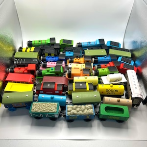 May include: A collection of colorful wooden toy trains and train cars. The trains are painted in various colors, including red, green, blue, and yellow. Some cars have cargo, and others have text like "Sodor Fuel."