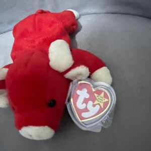 Puede incluir: Un juguete de peluche rojo con forma de toro, con detalles blancos en las pezuñas y la cara. El juguete tiene un ojo negro y una etiqueta en forma de corazón. La etiqueta muestra el texto "TY" y una estrella.