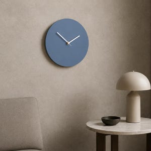May include: A circular wall clock with a matte blue face, featuring two thin white hands and a small gold-toned center cap. The clock has a clean, frameless design with no numbers or markings on the dial, providing a simple and functional timekeeping display.