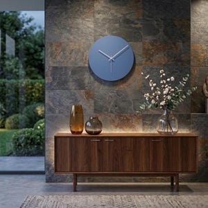 May include: A circular blue wall clock with thin white hands mounted on a dark stone wall above a wooden sideboard. The sideboard holds a tall amber glass vase and a shorter dark glass vase, alongside a clear glass vase filled with white flowers.