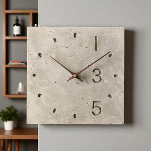 Könnte beinhalten: Quadratische Beton-Uhr mit braunen Zeigern und minimalistischem Design. Das Zifferblatt zeigt die Zahlen 1, 3 und 5, mit kleinen Punktmarkierungen. Eine moderne Wanduhr im industriellen Stil.