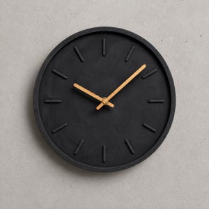 Könnte beinhalten: Eine runde Wanduhr mit einem mattschwarzen Zifferblatt und Rahmen sowie zwölf erhabenen schwarzen Stundenmarkierungen. Die Uhr hat zwei schlanke, helle Holzzeiger in der Mitte, die die Zeit mit einem schlichten Design anzeigen.