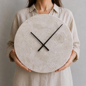 Könnte beinhalten: Eine runde Wanduhr mit einem strukturierten, hellbeigen, betonartigen Zifferblatt und zwei dünnen, schwarzen Metallzeigern. Das Zifferblatt weist eine raue, poröse Oberflächenbeschaffenheit mit natürlichen Farbvariationen auf, die mit den Zeigern kontrastieren.