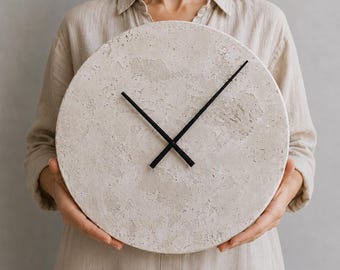 May include: A circular wall clock with a textured, light beige concrete-like face and two thin, black metal hands. The clock face features a rough, porous surface finish with natural variations in tone, contrasting with the sleek black hands.