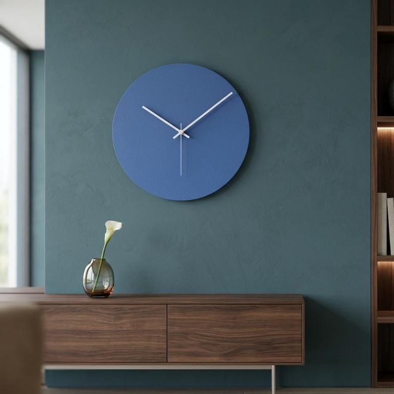 May include: A circular wall clock with a solid blue face and thin white hands. The clock features a simple, clean design without numbers or hour markers, mounted on a wall. The hands are positioned to indicate the time, with a central pin.