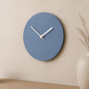 May include: A circular wall clock with a matte dusty blue face and two thin white hands. The center of the clock features a small gold-toned circular cap where the hands are attached. The design is simple, featuring no numbers or hour markers.