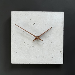 Könnte beinhalten: Eine quadratische Wanduhr mit einem hellgrauen Beton-Zifferblatt mit gesprenkelter, poröser Textur. Die Uhr hat zwei dünne, dunkelbraune Holzzeiger, die in der Mitte angebracht und mit einem kleinen, messingfarbenen Metallverschluss befestigt sind.