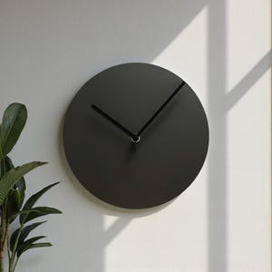 May include: A circular wall clock with a matte charcoal gray face and two thin black hands. The clock features a clean, solid design without any numbers or markings on the dial, providing a simple and functional timekeeping display.