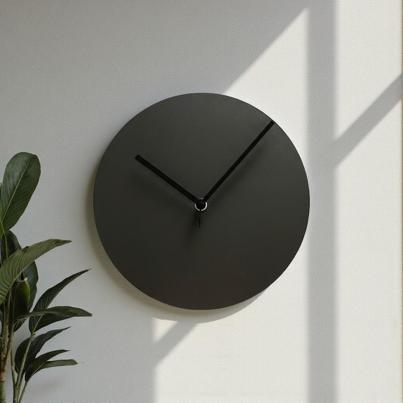 May include: A circular wall clock with a matte charcoal gray face and two thin black hands. The clock features a clean, solid design without any numbers or markings on the dial, providing a simple and functional timekeeping display.
