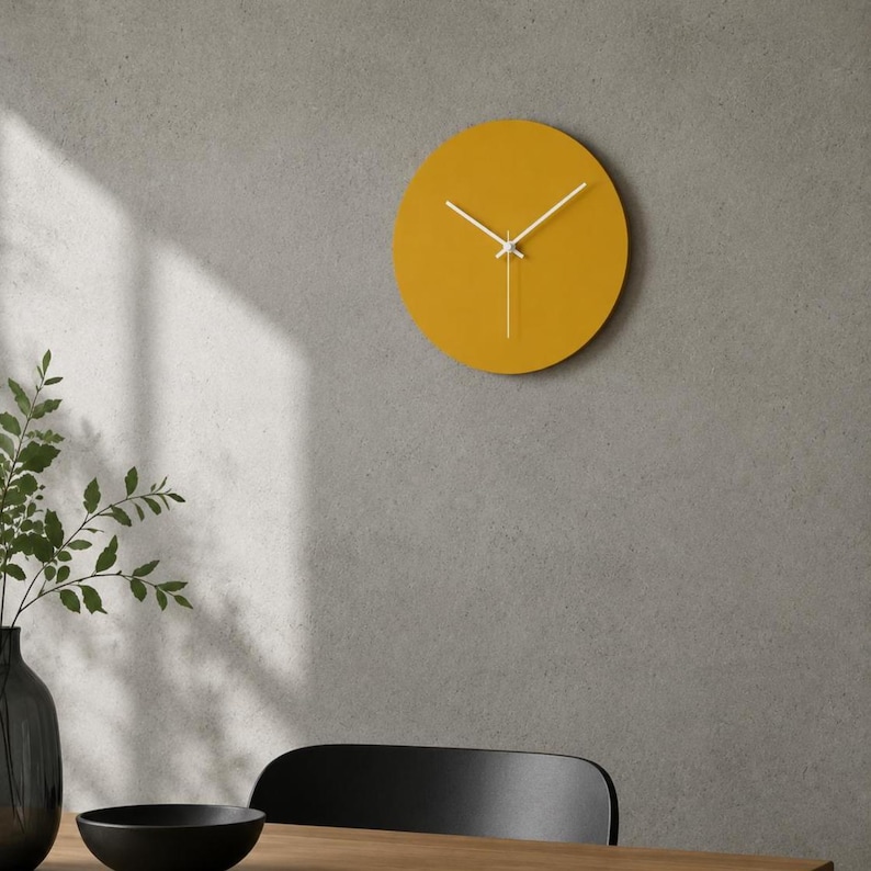 May include: A circular wall clock with a solid mustard yellow face and thin white hour, minute, and second hands. The clock features a smooth, flat surface without numbers or markers, providing a clean and simple aesthetic for any interior wall.