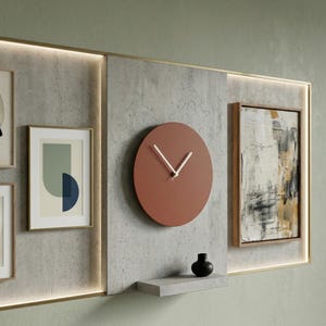 May include: A circular terracotta-colored wall clock with thin white hands mounted on a textured gray concrete panel. The panel features a small matching concrete shelf with a black vase, flanked by framed abstract art prints and integrated LED lighting.