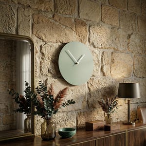 May include: A circular wall clock with a matte sage green face and two thin white hands. The clock is mounted on a textured stone wall, positioned above a wooden surface featuring a glass vase with dried foliage and a small green bowl.