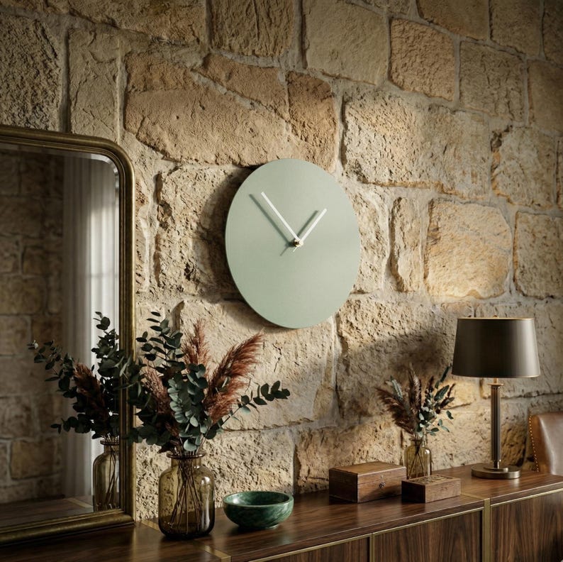 May include: A circular wall clock with a matte sage green face and two thin white hands. The clock is mounted on a textured stone wall, positioned above a wooden surface featuring a glass vase with dried foliage and a small green bowl.