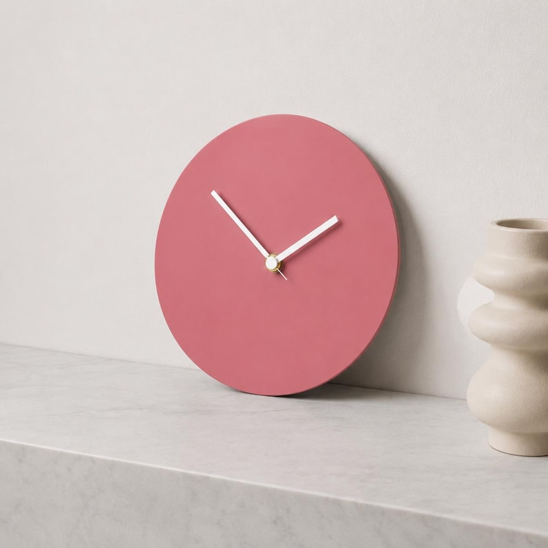 May include: A circular wall clock with a solid matte dusty rose face and two simple white hands. The clock features a small gold-toned center cap where the hands meet, providing a clean and functional design for tracking time in any room.