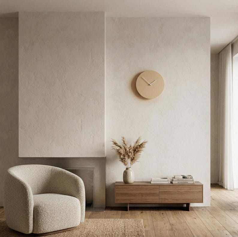 May include: A circular wall clock with a light beige face and thin white hands mounted on a textured off-white wall. The clock features a simple, clean design without numbers or markers, positioned above a wooden media console and a textured armchair.