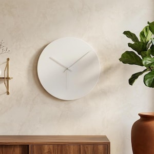 May include: A circular wall clock with a solid white face and thin, matching white hour and minute hands. The clock features a clean, smooth surface without any numbers or markings, providing a simple and functional timekeeping display.