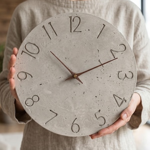 Könnte beinhalten: Eine runde Wanduhr aus hellgrauem Beton mit einer gesprenkelten, strukturierten Oberfläche. Die Uhr verfügt über vertiefte, dunkel gefärbte Ziffern von eins bis zwölf und zwei dünne, dunkelbraune Holzzeiger in der Mitte des Zifferblatts.