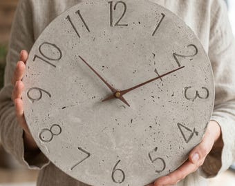 May include: A circular wall clock made of light gray concrete with a speckled, textured surface. The clock features recessed, dark-colored numerals from one to twelve and two thin, dark brown wooden hands centered on the face.