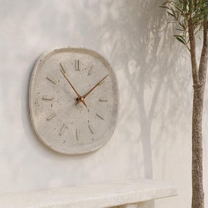 Könnte beinhalten: Eine runde Wanduhr mit einem gesprenkelten, hellgrauen Beton-Zifferblatt und einem erhabenen, strukturierten Rand. Die Uhr verfügt über zwölf kurze, rechteckige erhabene Stundenmarkierungen und zwei dünne, spitze braune Holzzeiger.
