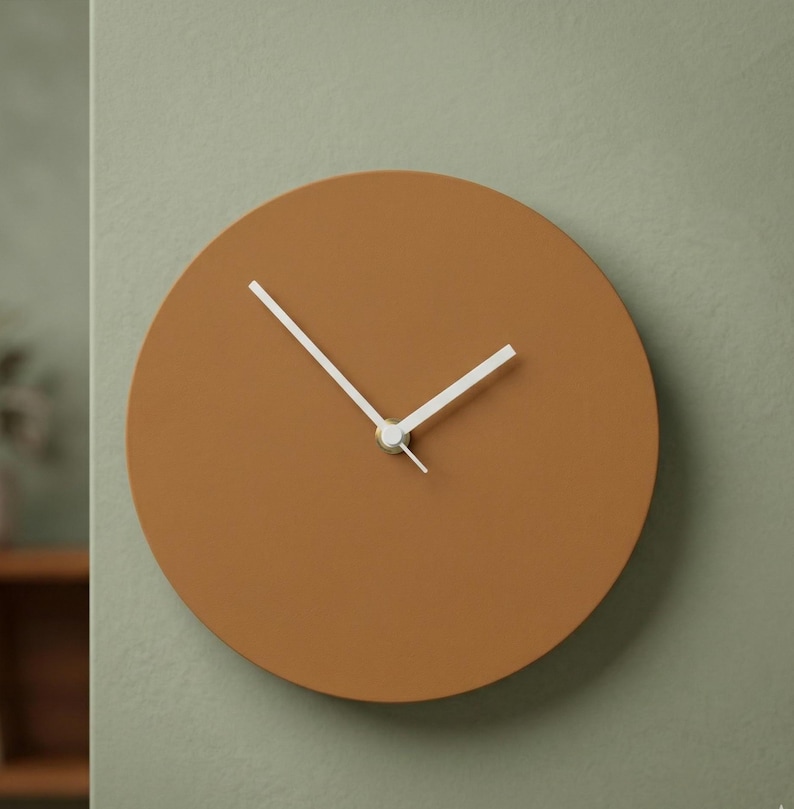 May include: A circular wall clock with a solid matte terracotta brown face and two simple white hands. The clock features a clean, frameless design with no numbers or markings on the dial, centered around a small silver-toned central hub.