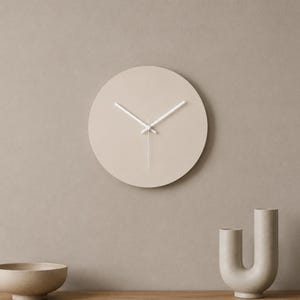 May include: A circular wall clock with a smooth, matte beige face and thin, white hour and minute hands. The clock features a clean, flat design without numbers or markers on the face, providing a simple and functional timekeeping display.