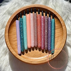 May include: A collection of 14 glitter pens in various colors, including purple, teal, pink, and orange, arranged in a wooden bowl. The pens have black caps and are all the same size.