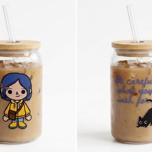 May include: Glass tumbler with a bamboo lid and straw, filled with iced coffee. One tumbler features a cartoon character in a yellow jacket. The other tumbler has the text "Be careful what you wish for" and a black cat.