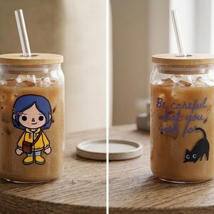 May include: Two glass tumblers filled with iced coffee, bamboo lids, and glass straws. One has a cartoon character, the other the text "Be careful what you wish for" and a black cat. Both are on a wooden table.