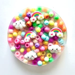 May include: A circular resin coaster filled with colourful beads in shades of pink, yellow, orange, green, and blue. The coaster features small, smiling white cloud charms and rainbow charms with heart accents.