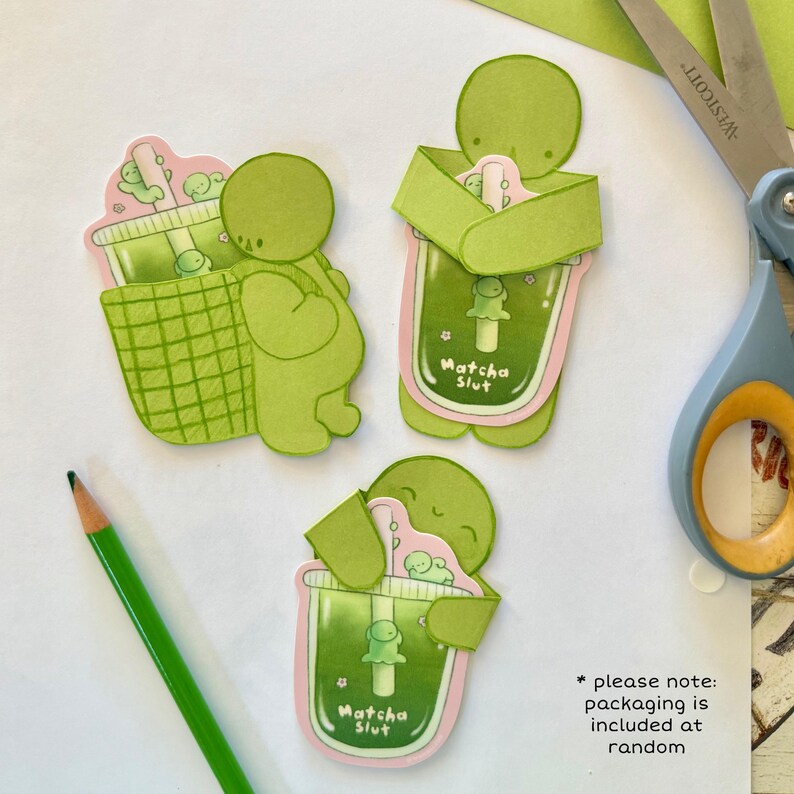May include: Green and pink die-cut stickers featuring a cartoon character holding a matcha drink. The stickers have the text "Matcha Slut" and are placed on a white surface with a green pencil and scissors.