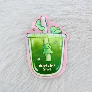May include: A sticker depicting a green matcha drink with a straw and three small green figures. The drink is in a pink-edged cup with the words "Matcha Slut" in white script. Small pink flowers are scattered around the cup.