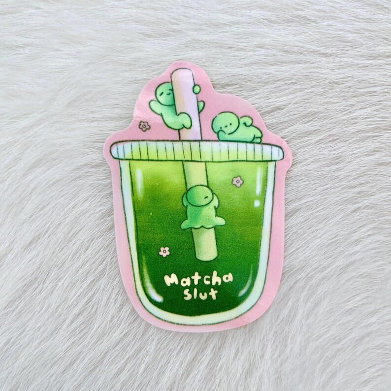 May include: A sticker depicting a green matcha drink with a straw and three small green figures. The drink is in a pink-edged cup with the words "Matcha Slut" in white script. Small pink flowers are scattered around the cup.