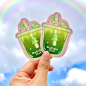 May include: Two stickers featuring matcha drinks with green liquid and a straw. The stickers have a pink border and the text "Matcha Slut". The drinks have a cute green creature on top and a flower. The background is a blue sky with a rainbow.