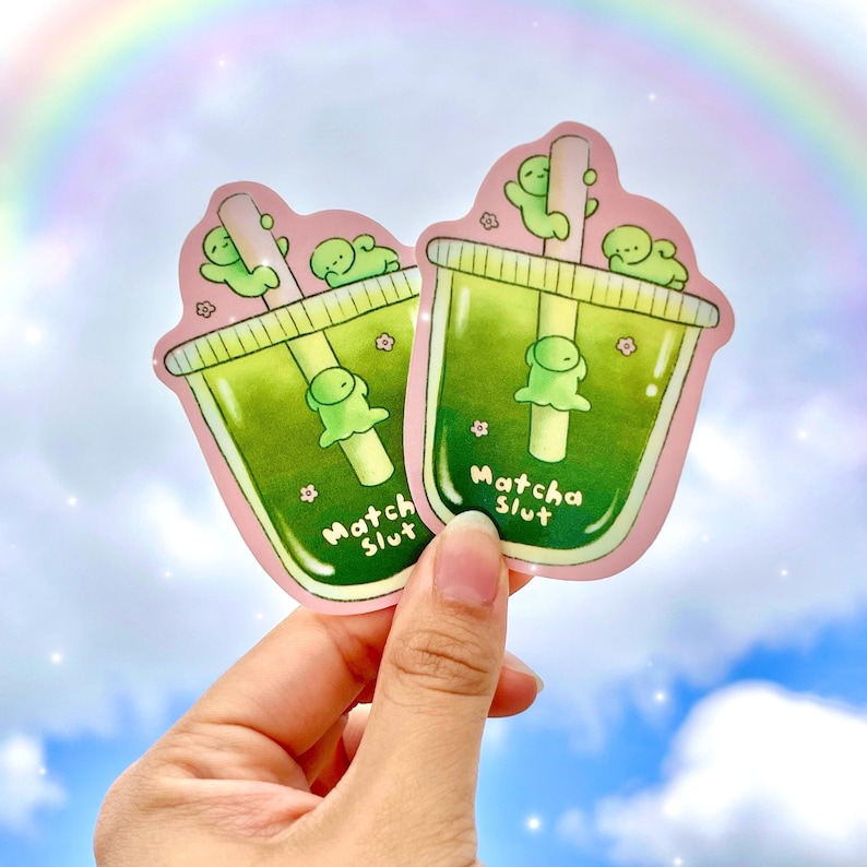 May include: Two stickers featuring matcha drinks with green liquid and a straw. The stickers have a pink border and the text "Matcha Slut". The drinks have a cute green creature on top and a flower. The background is a blue sky with a rainbow.