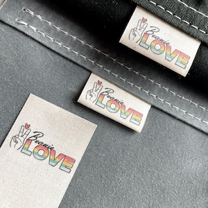 May include: Three white fabric labels with a rainbow colored "Because Love" text and a peace sign with a red heart.
