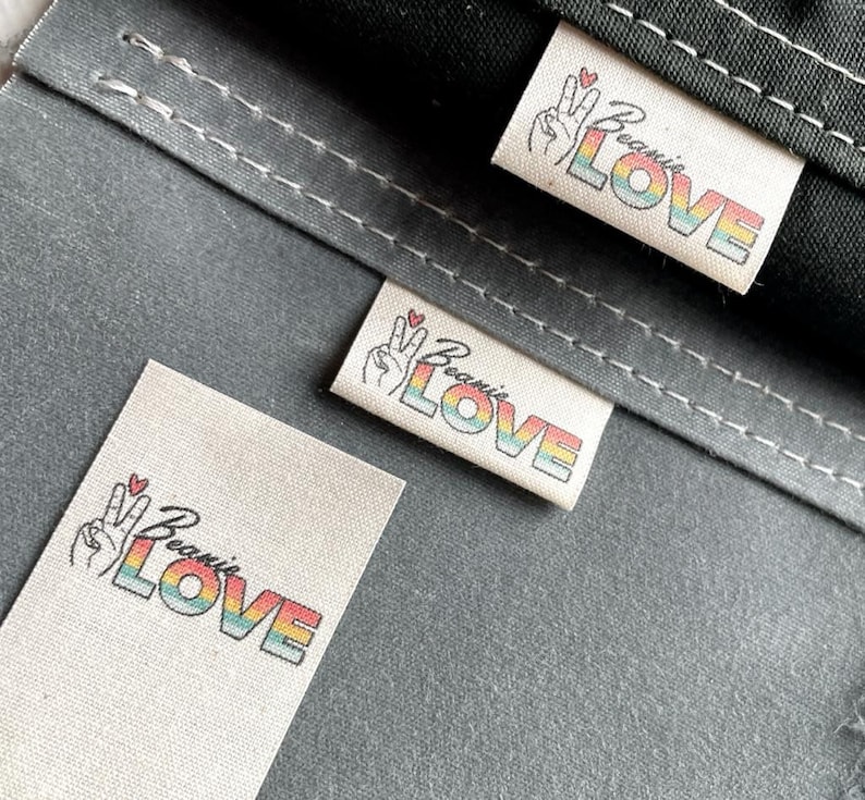 May include: Three white fabric labels with a rainbow colored "Because Love" text and a peace sign with a red heart.